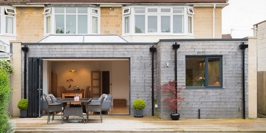 Flat-roof extensions with bi-fold doors, open onto timber patio with dining table and chairs.